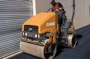 A worker operates a yellow CASE asphalt roller, smoothing freshly paved blacktop beside a building with a metal roll-up door.