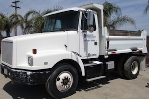 A white dump truck with a clean cab and open cargo bed, parked outdoors under a sunny sky with palm trees in the background.