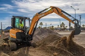 A construction worker operates an orange CASE excavator, digging into a dirt mound at a sunny outdoor worksite.