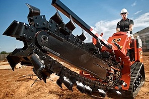 A construction worker operates a large orange trencher with a massive toothed chain, digging into the dirt at a worksite.